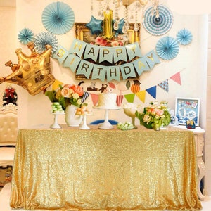 May include: A festive birthday party scene with a gold sequin tablecloth. Decorations include a "Happy Birthday" banner, balloons, flowers, and a cake. The scene is set for a celebration with a crown balloon and other party decorations.