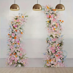 May include: A floral arch and pillar arrangement with a variety of pastel-colored flowers, including pink, white, and orange blooms. The arrangement is set against a white wall with three gold pendant lights hanging above.