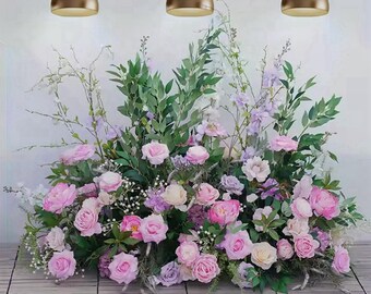 May include: A floral arrangement featuring pink roses, lavender flowers, and green foliage. The flowers are arranged on a wooden surface, with three gold pendant lights hanging above. The overall aesthetic is soft and romantic.