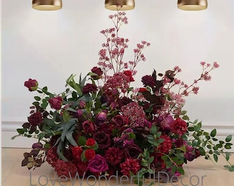 May include: A large floral arrangement with deep red and burgundy roses, peonies, and other flowers. The arrangement is in a clear vase and is sitting on a wooden floor. There are three gold pendant lights hanging above the arrangement.