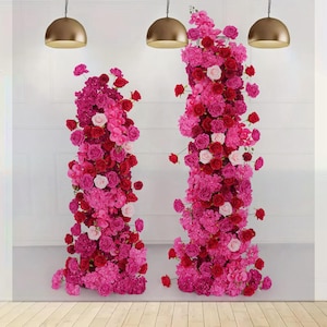 Op de afbeelding: Twee verticale bloemstukken met rode, roze en witte rozen en hortensia's. Ze staan tegen een witte muur. Gouden hanglampen hangen erboven. Een kleurrijke bloemenpracht.