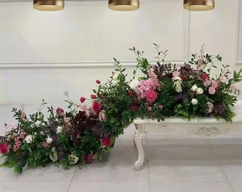 May include: A white table with ornate legs is adorned with a lush floral arrangement. The arrangement features a variety of flowers in shades of pink, red, and white, complemented by green foliage. Three bronze pendant lights hang above.