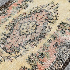 May include: Vintage-style rug with a distressed look. The rug features a central floral design in shades of peach, green, and black on a cream background. The border is a dark brown with a repeating pattern. The rug has a worn appearance.