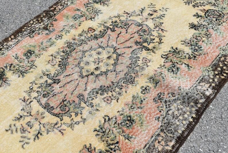 May include: Vintage-style rug with a distressed look. The rug features a central floral design in shades of peach, green, and black on a cream background. The border is a dark brown with a repeating pattern. The rug has a worn appearance.
