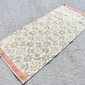 May include: Rectangular rug with a floral pattern in cream, orange, and green. The rug has a distressed look with an orange border. The design features repeating floral medallions and other floral elements. This rug is suitable for home decor.