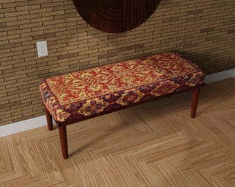 May include: A rectangular bench with a patterned red, gold, and blue upholstered top. The bench has four dark brown wooden legs. The pattern on the top resembles a traditional rug design. The bench is placed on a wooden floor.