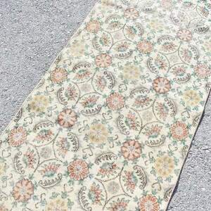 May include: A rectangular rug with a floral pattern. The rug features a cream-colored background with repeating floral designs in shades of orange, yellow, and green. The rug's edges have a reddish-orange border. The rug is placed on a gray surface.