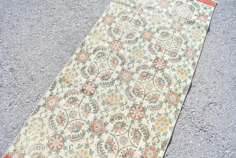 May include: A rectangular rug with a floral pattern. The rug features a cream-colored background with repeating floral designs in shades of orange, yellow, and green. The rug's edges have a reddish-orange border. The rug is placed on a gray surface.