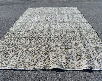 May include: A rectangular, beige rug with an intricate floral pattern in dark green and black. The rug is laid out on a dark gray surface, showcasing its full length and design. The rug's edges are slightly frayed.