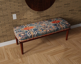 May include: A rectangular bench with a patterned cushion and wooden legs. The cushion features a blue background with orange and cream-colored geometric designs. The bench is set against a brick wall and wooden floor.