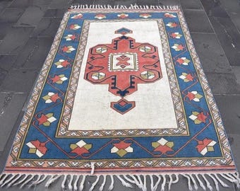 May include: Rectangular rug with a central geometric design in red, blue, and cream. The rug features a blue border with floral and geometric patterns, and white fringe along the edges. The rug is placed on a dark gray stone surface.