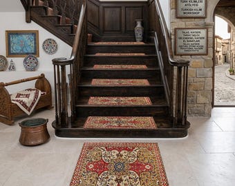 May include: A red and beige patterned rug sits on a light-colored floor in front of a dark wooden staircase. Stair treads feature matching rugs. The staircase has a dark wooden banister and is decorated with framed artwork and a vase.