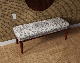 May include: A rectangular bench with a patterned cushion and dark wooden legs. The cushion features a white background with a floral and medallion design in shades of gray and blue. The bench is set against a brick wall and wooden floor.
