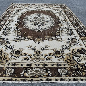 May include: A vintage rug with a brown and white floral pattern. The rug has a central medallion with a geometric design. The rug is made of wool and has a fringe border.