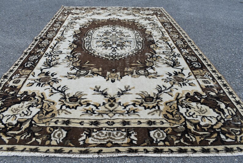 May include: A vintage rug with a brown and white floral pattern. The rug has a central medallion with a geometric design. The rug is made of wool and has a fringe border.