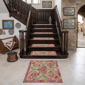 May include: A detailed shot of a staircase adorned with Turkish Kilim rugs. The rugs feature floral patterns in pink, red, and green on a beige background. The staircase is made of dark wood, and the walls display decorative plates and signs with Turkish text.