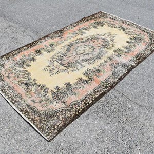 May include: An antique area rug with a central floral medallion design. The rug features a cream-colored field with a pink and black border. The rug's design includes floral and botanical elements, creating a vintage aesthetic.