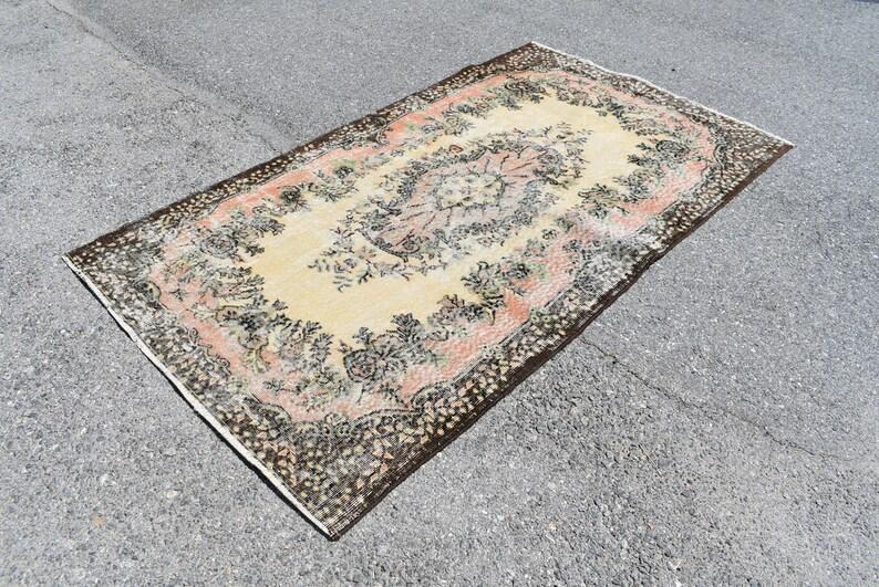 May include: An antique area rug with a central floral medallion design. The rug features a cream-colored field with a pink and black border. The rug's design includes floral and botanical elements, creating a vintage aesthetic.
