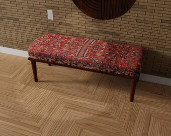 May include: A rectangular bench with a patterned red and blue upholstered top. The bench has dark brown wooden legs and frame. The patterned top features a traditional rug design. The bench is placed on a light brown wooden floor.