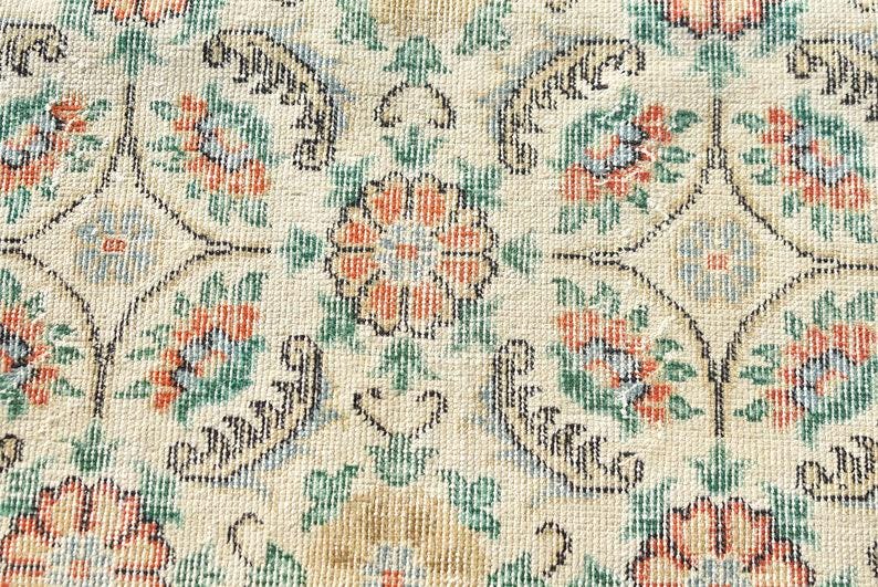 May include: Close-up of a beige rug with a floral pattern. The design features orange, green, and blue flowers, with black decorative elements. The rug has a vintage, distressed look.