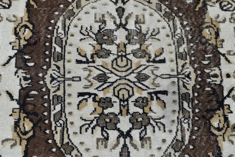 May include: A close-up of a beige rug with a brown border. The rug has a repeating pattern of stylized flowers and leaves in black, brown, and beige.