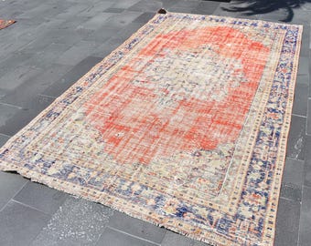 May include: A rectangular area rug featuring a large central orange field with a faded cream and blue medallion pattern. The rug has a wide, intricate border in deep blue and cream tones, with short fringe detailing along the short edges.