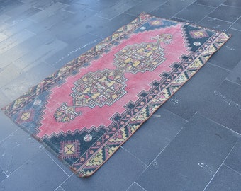 May include: A rectangular, vintage rug with a central pink field and a detailed geometric pattern in yellow, black, and cream. The rug has a dark blue border and is likely made of wool.