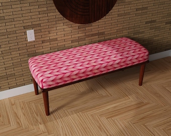 May include: A rectangular bench with a pink and white woven pattern seat. The bench has a dark brown wooden frame and four legs. The bench is placed on a wooden floor.