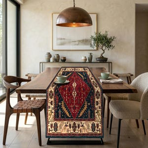 May include: A rectangular table runner with an intricate, traditional rug design. The runner features a central red diamond pattern, framed by navy blue and beige borders. The runner is displayed on a wooden table, with chairs and decor in the background.