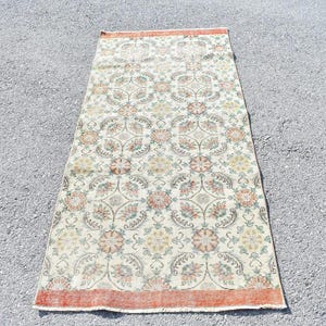 May include: Rectangular vintage rug with a floral pattern in cream, orange, and green. The rug has a distressed look with a faded design. The border is a darker orange. The rug is likely made of wool or a similar material.