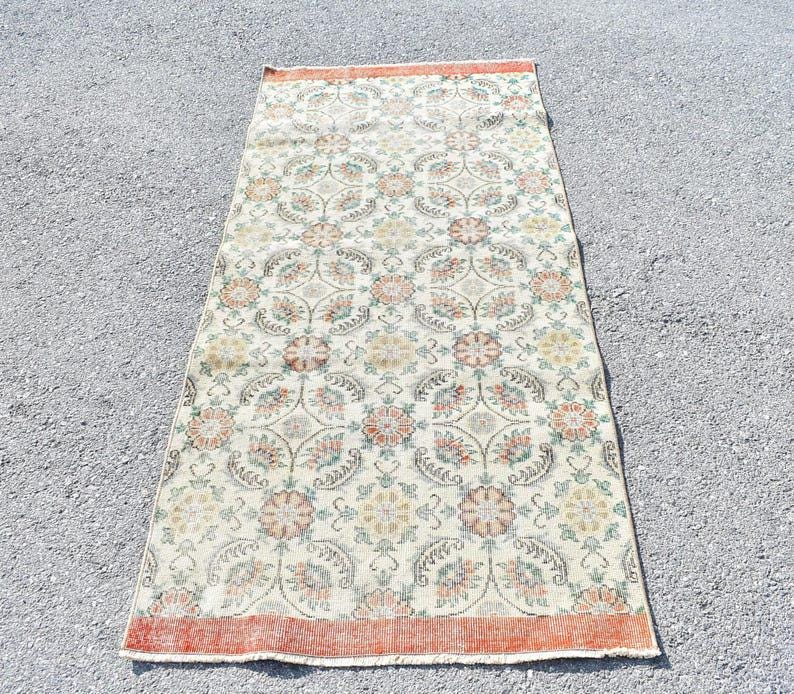 May include: Rectangular vintage rug with a floral pattern in cream, orange, and green. The rug has a distressed look with a faded design. The border is a darker orange. The rug is likely made of wool or a similar material.