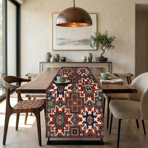May include: A rectangular table runner with a geometric pattern in red, blue, cream, and green. The runner is placed on a wooden dining table, with chairs and a copper pendant light visible in the background. The runner's design features star-like and geometric shapes.