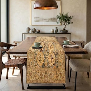 May include: A rectangular table runner featuring an intricate, traditional-style pattern in shades of gold, beige, and muted teal. The textile displays a dense, symmetrical floral and vine motif with a decorative border, centered on a wooden table.