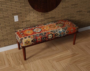 May include: A rectangular bench with a patterned top featuring a mix of geometric shapes in red, orange, blue, and yellow. The bench has dark brown wooden legs and a matching frame. The bench is placed on a wooden floor.