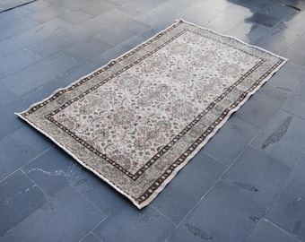 Könnte beinhalten: Rechteckiger beiger Teppich mit floralem Muster und dunkelbraunem Rand. Der Teppich liegt auf einer dunkelgrauen Fliesenoberfläche. Das Design des Teppichs zeigt ein zentrales Blumenmotiv und einen detaillierten Rand.