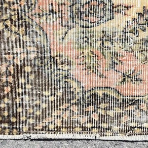 May include: Close-up of a vintage rug with a distressed design. The rug features a floral pattern in shades of peach, cream, and green, with a dark brown border. The rug has a worn, antique appearance, suggesting it is an older piece.