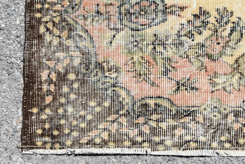 May include: Close-up of a vintage rug with a distressed design. The rug features a floral pattern in shades of peach, cream, and green, with a dark brown border. The rug has a worn, antique appearance, suggesting it is an older piece.