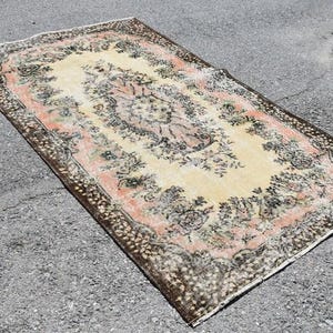 May include: Rectangular rug with a central floral medallion in cream, surrounded by a pink border and a dark brown patterned edge. The rug has an aged, vintage appearance, with faded colors and a distressed look.