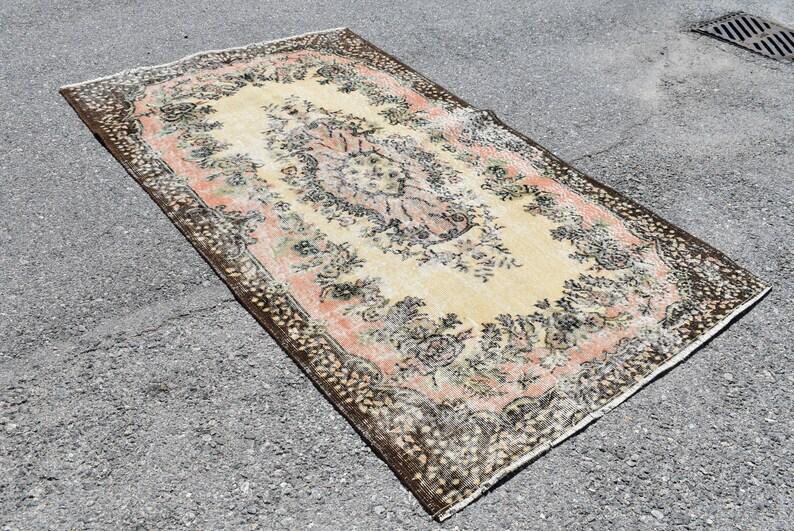 May include: Rectangular rug with a central floral medallion in cream, surrounded by a pink border and a dark brown patterned edge. The rug has an aged, vintage appearance, with faded colors and a distressed look.