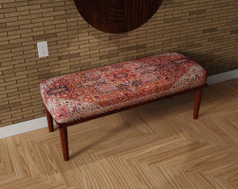 May include: A rectangular bench with a patterned, upholstered seat in shades of red, orange, and cream. The bench has a dark brown wooden frame and four short, rounded legs. The bench is placed on a wooden floor.