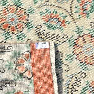 May include: Close-up of a beige rug with a floral pattern in orange, green, and blue. The rug has a distressed look, with a section folded over, revealing a red border and a tag that reads "RLO 1572".