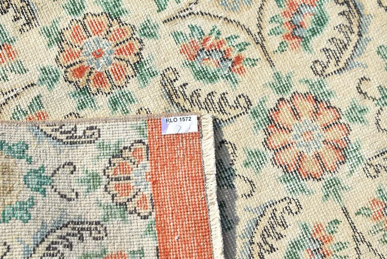 May include: Close-up of a beige rug with a floral pattern in orange, green, and blue. The rug has a distressed look, with a section folded over, revealing a red border and a tag that reads "RLO 1572".