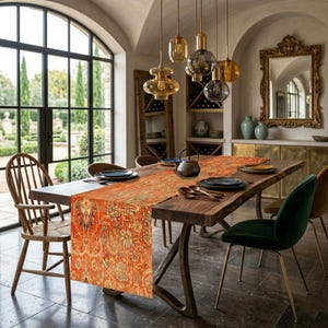 May include: A dining room table with a long, ornate orange and beige patterned table runner. The table is made of dark wood and has metal legs. There are plates, utensils, and a teapot on the table. The room has large windows and decorative lighting.