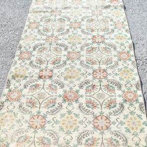 May include: A rectangular, floral patterned rug with a cream-colored background. The rug features a repeating pattern of orange, yellow, and green flowers and leaves. The rug is placed on a gray surface.
