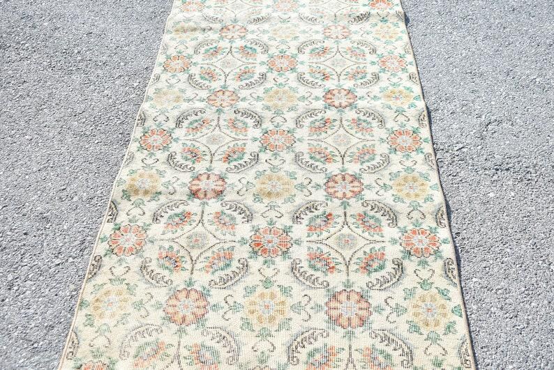 May include: A rectangular, floral patterned rug with a cream-colored background. The rug features a repeating pattern of orange, yellow, and green flowers and leaves. The rug is placed on a gray surface.