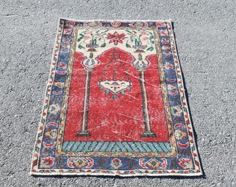 May include: A small, rectangular rug with a distressed, vintage appearance. The rug features a red central field with a central design and a border of blue, cream, and floral patterns. The rug is likely hand-knotted.