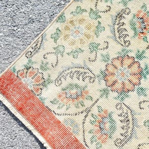 May include: Close-up of a section of a vintage rug with a cream-colored background. The rug features floral patterns in shades of orange, green, and blue, with a red border. The design is intricate and detailed, showcasing a classic aesthetic.