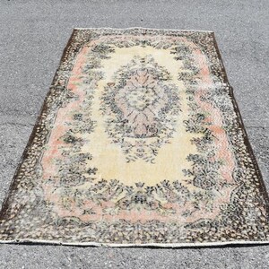 May include: Rectangular rug with a central floral medallion in cream, surrounded by a border of pink and black floral patterns. The rug has a distressed, vintage appearance, with a dark brown border.