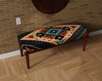 May include: A wooden bench with a rectangular seat featuring a colorful geometric pattern in shades of teal, red, beige, and black. The bench has four wooden legs and is placed on a wooden floor.