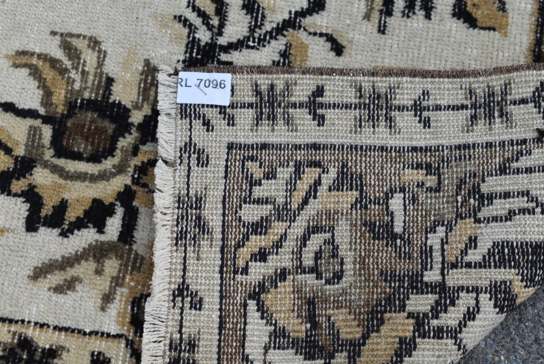 May include: Close-up of a beige rug with a brown and black floral pattern. The rug has a fringe border and a label that reads "RL 7096".